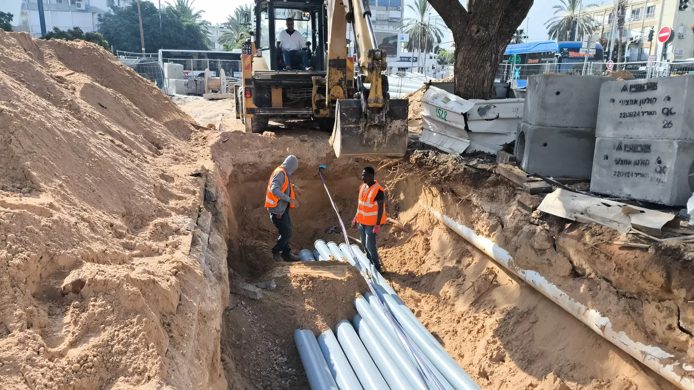 תעלה פתוחה לאורך הרחוב במקטע G1-5-3: שורת צינורות מונחת בקרקעית, שני עובדים בחפירה, מחפרון ואלמנטי בטון טרום ערוכים בצד.