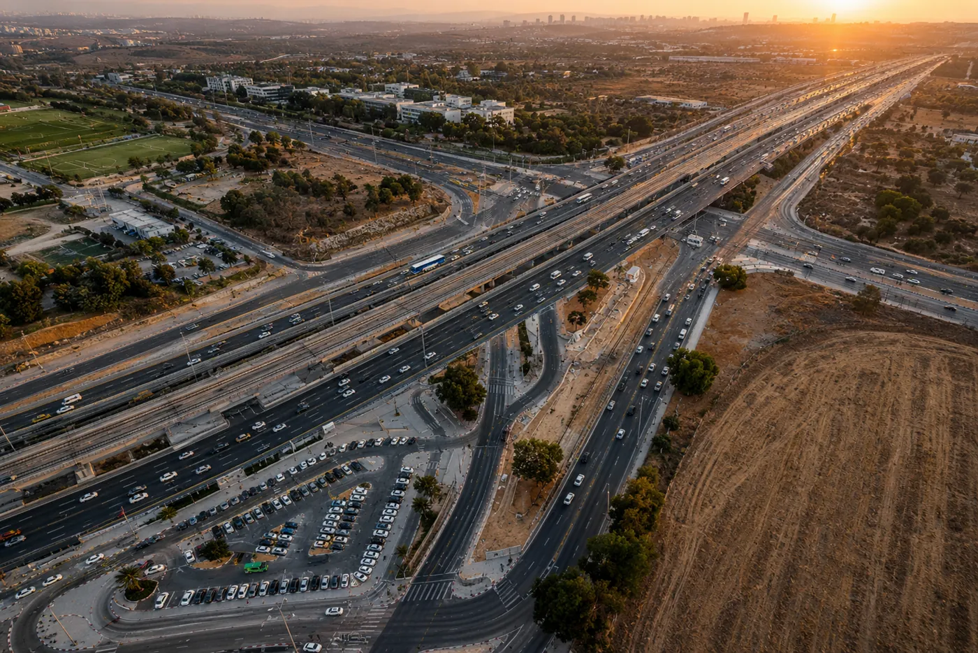 צילום אוויר בשעת שקיעה על מחלף גדול: נתיבי הכביש, מסילות הרכבת הקלה לצדם ותנועה ערה, עם קו הרקיע של העיר באופק.