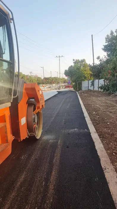 סלילת אספלט והידוק במכבש לאורך המדרכה.