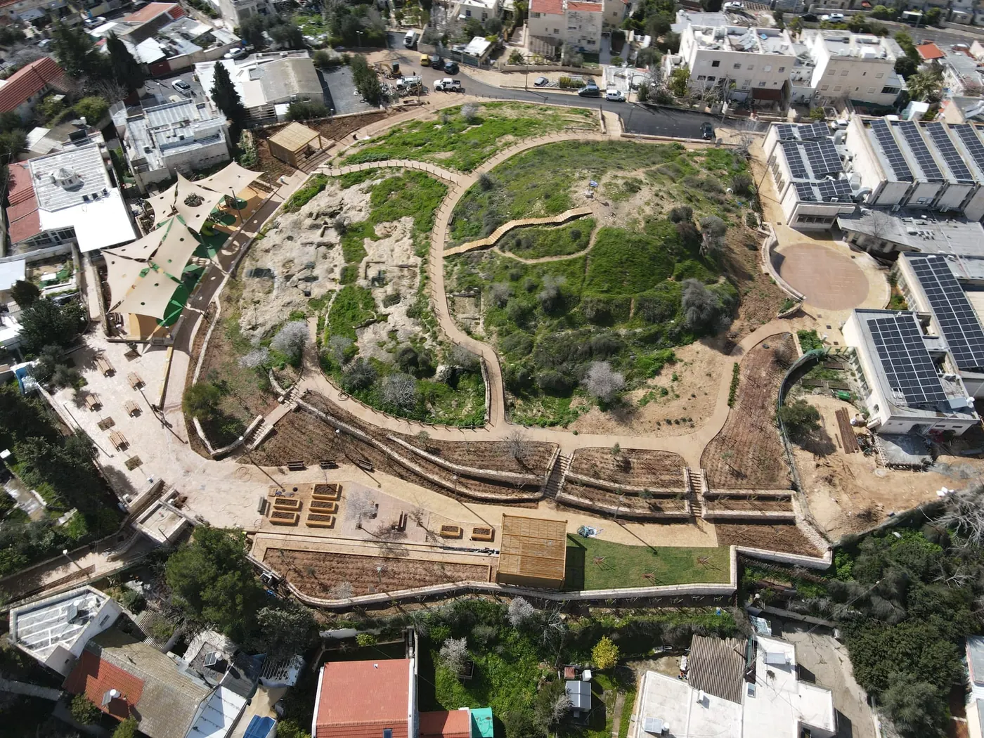 צילום אוויר של פארק הרוגם בירושלים: גבעה מגוננת עם שבילים מתפתלים, קירות תמך מדורגים ומדרגות ישיבה מעץ, בלב שכונת מגורים.