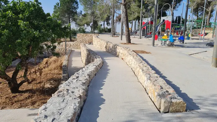 שביל מוגמר בפארק: קיר אבן נמוך לאורכו, עצים בוגרים שנשמרו ומתקני משחק ברקע.