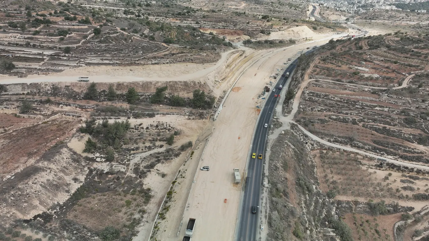 צילום רחפן של מקטע בכביש 60 בביצוע: הכביש הקיים עם תנועה שוטפת, ולצדו תוואי רחב חדש בעבודות עפר, קירות תומכים ומעבירי מים, בין מדרונות חקלאיים מדורגים.